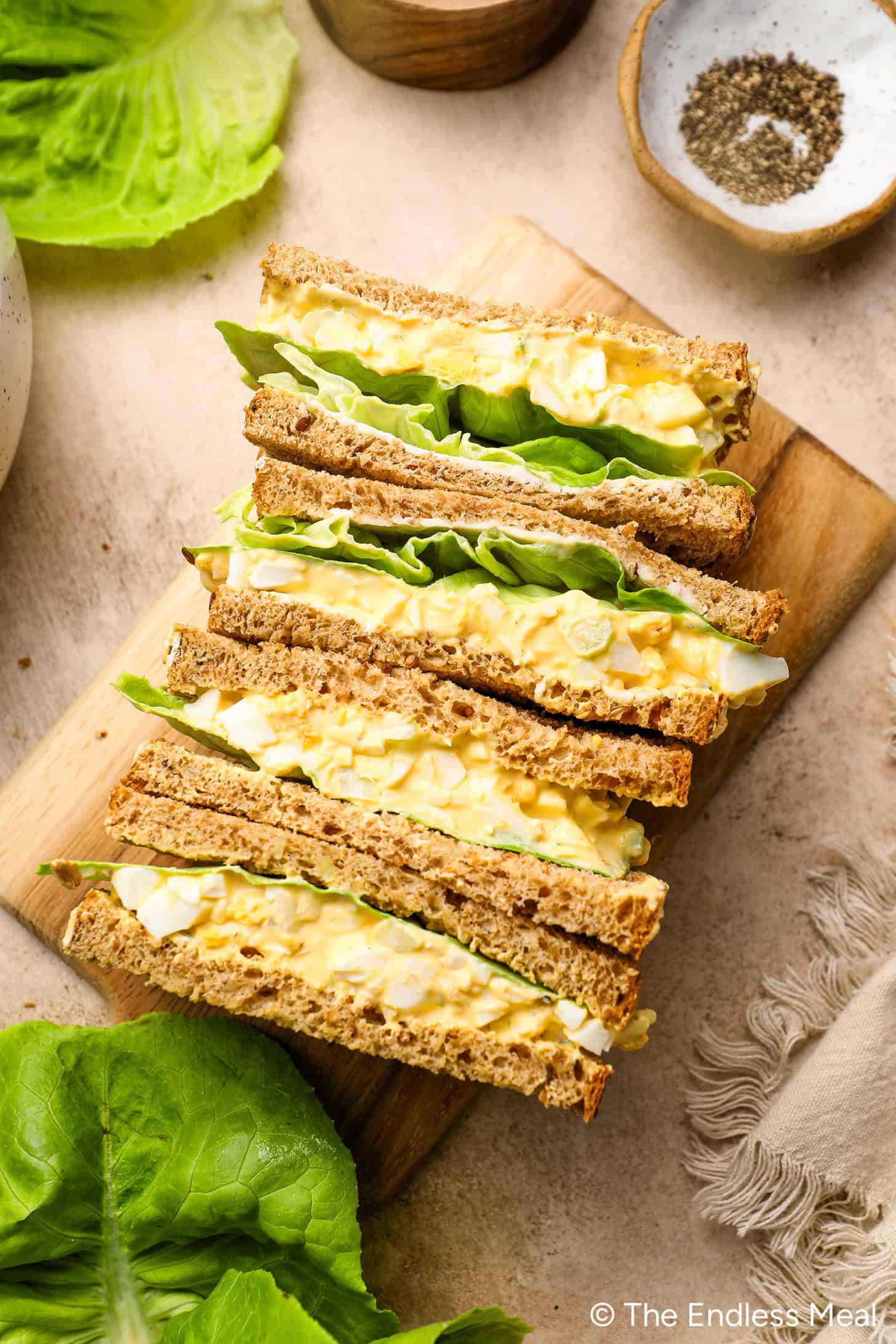 Four egg salad sandwiches with lettuce on whole wheat bread, stacked on a wooden board. A bowl of black pepper and lettuce leaves are nearby.