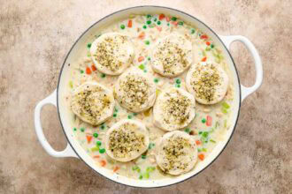 A white casserole dish filled with creamy Chicken Pot Pie with Biscuits and vegetables, topped with eight herb-seasoned biscuits, sits on a brown countertop.