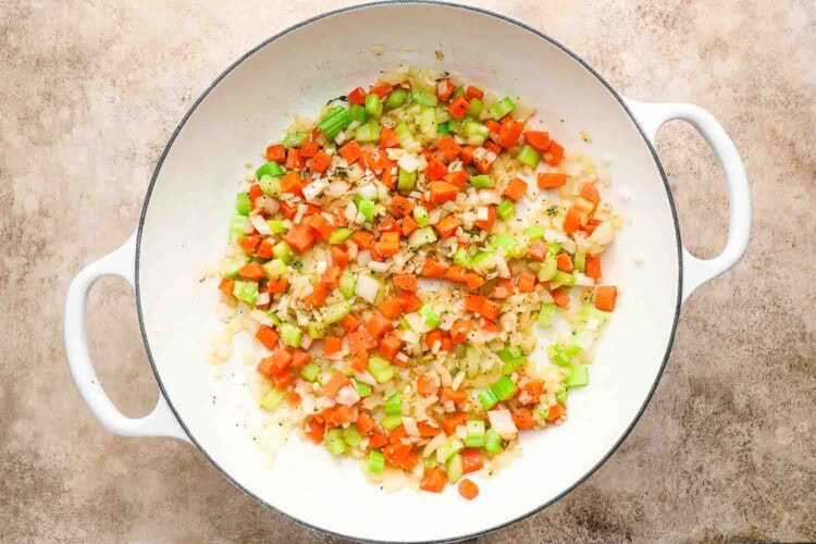 Chopped onions, carrots, and celery sautéing in a white pot on a beige surface—the perfect base for a comforting Chicken Pot Pie with Biscuits.