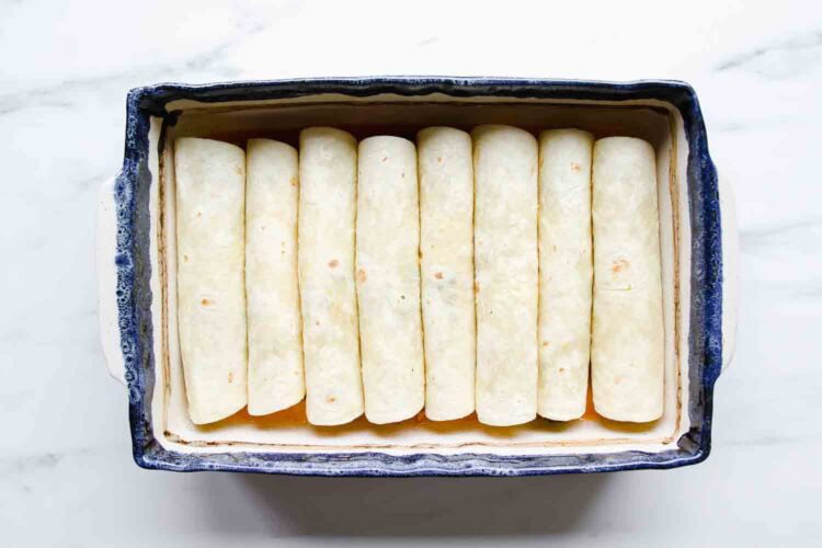 A rectangular baking dish containing ten rolled Chicken Enchiladas arranged in a single layer on a white surface.