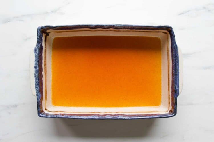 A rectangular baking dish filled with an orange liquid, perfect for making Chicken Veggie Enchiladas, sits on a white marble surface.