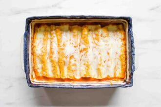 Rectangular baking dish filled with cheese-topped Chicken Enchiladas in red sauce, viewed from above on a white surface.