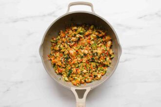 A beige skillet contains a cooked mixture of diced vegetables and herbs for veggie enchiladas, displayed on a white marble surface.