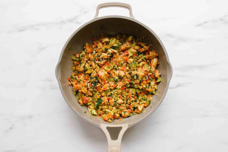 A beige skillet contains a cooked mixture of diced vegetables and herbs for veggie enchiladas, displayed on a white marble surface.