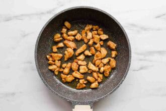 Cooked pieces of seasoned chicken breast for Chicken Veggie Enchiladas in a black nonstick frying pan on a white marble surface.