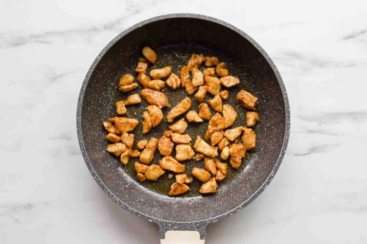 Cooked pieces of seasoned chicken breast for Chicken Veggie Enchiladas in a black nonstick frying pan on a white marble surface.