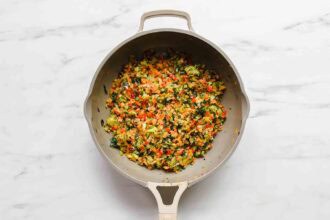 A sauté pan filled with chopped and cooked mixed vegetables, perfect for veggie enchiladas, including carrots, bell peppers, and leafy greens, on a marble countertop.