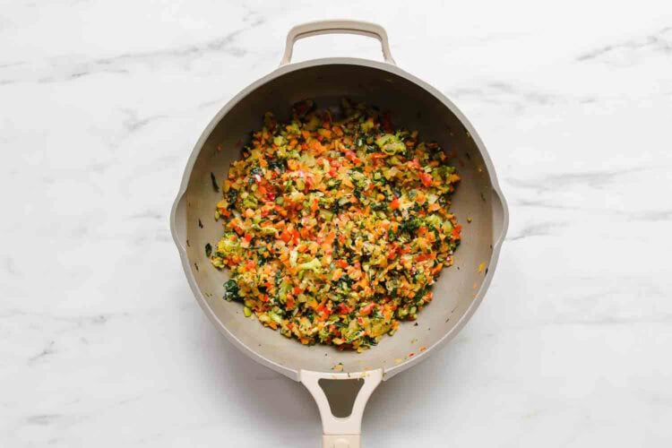 A sauté pan filled with chopped and cooked mixed vegetables, perfect for veggie enchiladas, including carrots, bell peppers, and leafy greens, on a marble countertop.
