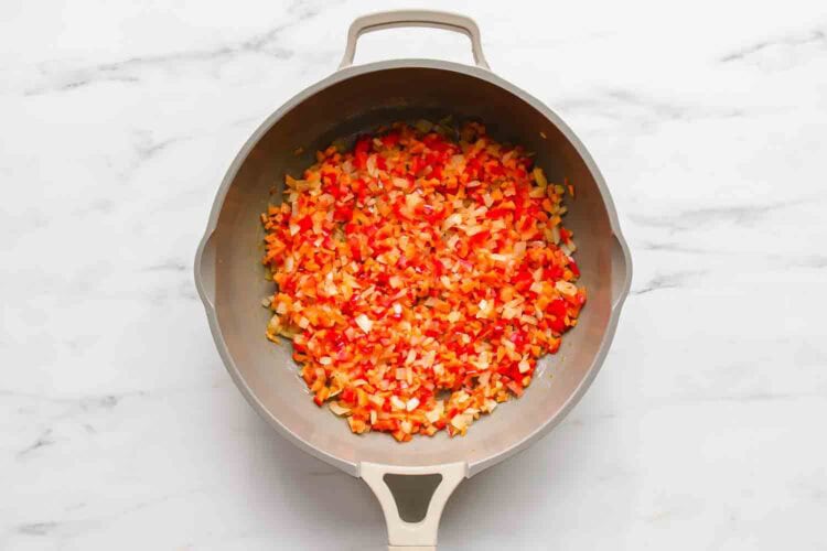 A frying pan on a white surface contains finely diced red and yellow bell peppers and onions being sautéed, perfect as a flavorful base for Chicken Veggie Enchiladas.