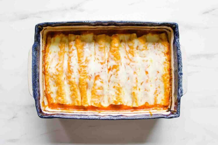 Rectangular baking dish filled with cheese-topped Chicken Enchiladas in red sauce, viewed from above on a white surface.