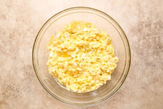 A glass bowl filled with egg salad mixture, featuring chopped eggs and mayonnaise, placed on a light-colored surface.