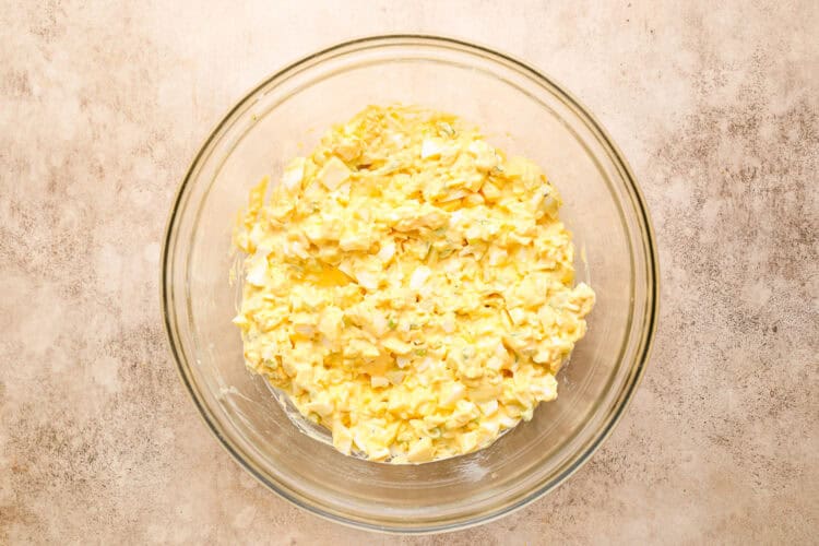 A glass bowl filled with egg salad mixture, featuring chopped eggs and mayonnaise, placed on a light-colored surface.