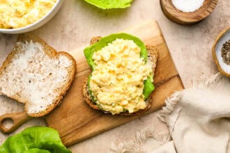 An open-faced egg salad sandwich on whole grain bread with a lettuce leaf on a wooden board, surrounded by a bowl of egg salad, mayonnaise, and seasonings.