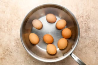 A stainless steel saucepan containing eight brown eggs sits on a beige textured surface.