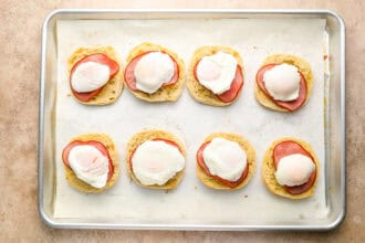 A baking sheet with eight halves of English muffins, each topped with ham and a poached egg, arranged in two rows on parchment paper—perfect for preparing homemade Eggs Benedict.