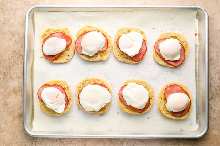 A baking sheet with eight halves of English muffins, each topped with ham and a poached egg, arranged in two rows on parchment paper—perfect for preparing homemade Eggs Benedict.