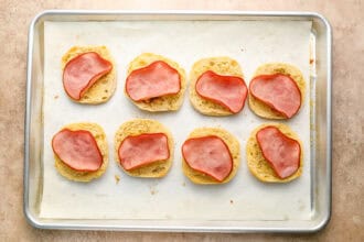 Eight biscuit halves topped with slices of ham are arranged on a parchment-lined baking sheet, ready to be transformed into homemade Eggs Benedict with rich, creamy Hollandaise sauce.