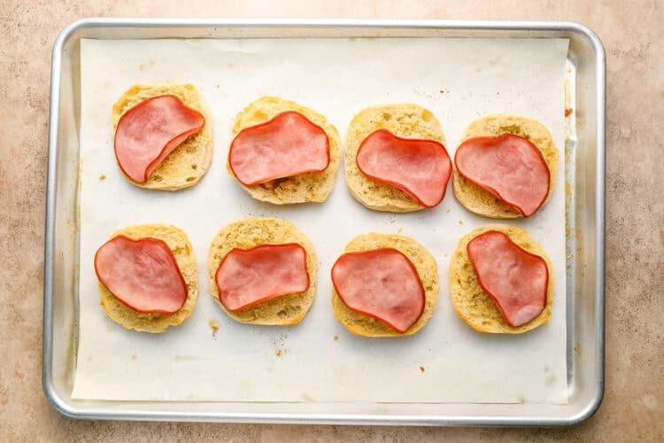 Eight biscuit halves topped with slices of ham are arranged on a parchment-lined baking sheet, ready to be transformed into homemade Eggs Benedict with rich, creamy Hollandaise sauce.