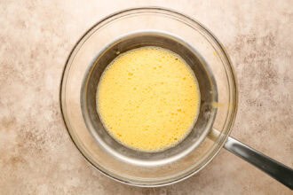 A metal saucepan with whisked yellow egg mixture, perfect for homemade Hollandaise sauce, sits inside a larger glass bowl on a light-colored countertop—an essential step in preparing classic breakfast favorites like Eggs Benedict.