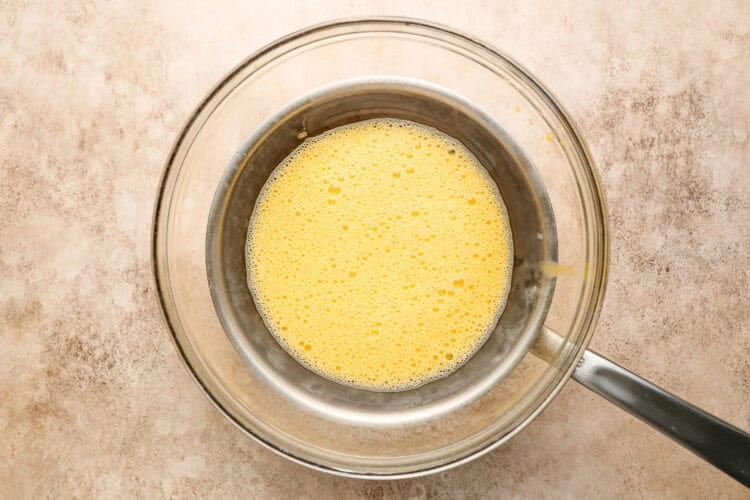 A metal saucepan with whisked yellow egg mixture, perfect for homemade Hollandaise sauce, sits inside a larger glass bowl on a light-colored countertop—an essential step in preparing classic breakfast favorites like Eggs Benedict.