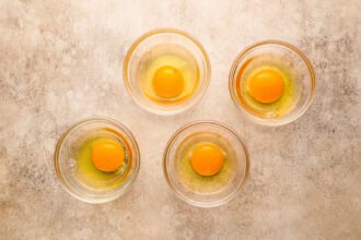 Four raw eggs in separate clear glass bowls are arranged on a light-colored textured surface, ready to be used for homemade Hollandaise sauce or the perfect Eggs Benedict.