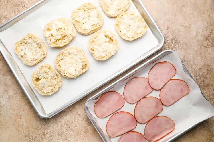 Two baking trays: one with six open biscuit halves, the other with eight slices of ham, perfect for assembling homemade Eggs Benedict on parchment paper atop a light-colored surface.