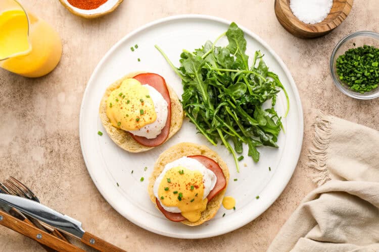 A plate with Classic Eggs Benedict on English muffins, topped with homemade hollandaise sauce and chives, served with a side of arugula.
