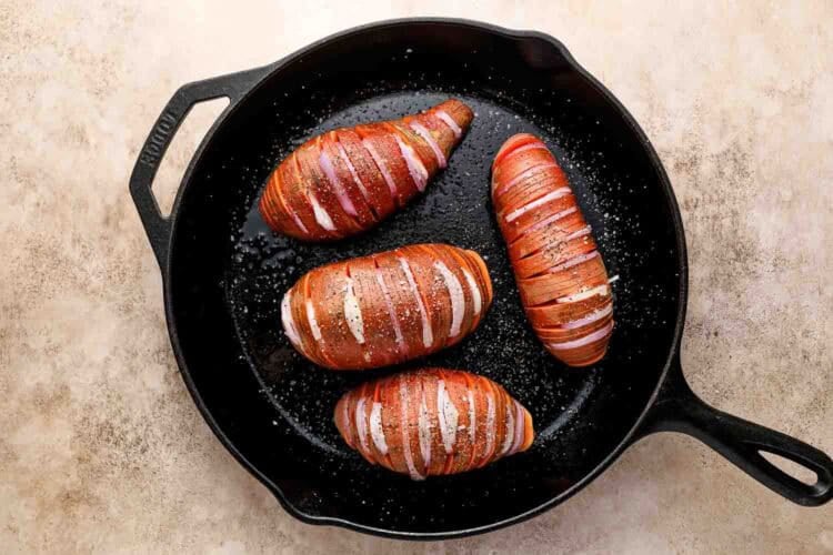 Four whole Hasselbacked Sweet Potatoes, sliced but not all the way through, rest in a black cast iron skillet and are sprinkled with salt and pepper, ready for baking.