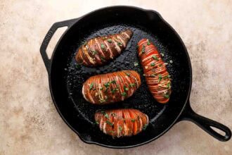 Three sliced Hasselback sweet potatoes garnished with herbs and garlic, cooked in a black cast iron skillet on a light-colored surface.