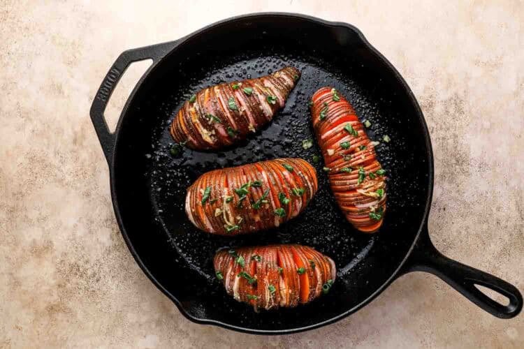 Three sliced Hasselback sweet potatoes garnished with herbs and garlic, cooked in a black cast iron skillet on a light-colored surface.