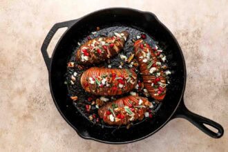 Four Hasselback Sweet Potatoes topped with cheese, nuts, and pomegranate seeds are arranged in a black cast iron skillet on a light surface.
