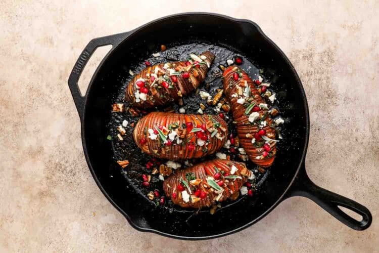 Four Hasselback Sweet Potatoes topped with cheese, nuts, and pomegranate seeds are arranged in a black cast iron skillet on a light surface.