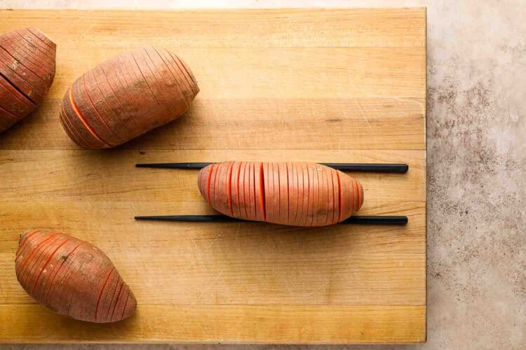 Three Hasselback Sweet Potatoes rest on a wooden cutting board, with one sweet potato placed between two chopsticks and sliced thinly but not all the way through.