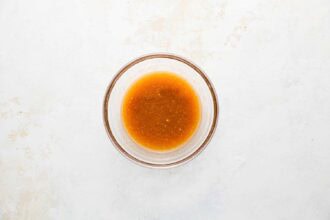 A clear glass bowl contains a small amount of light brown liquid, resembling a sauce or dressing for Holiday Brussels Sprouts, placed on a white textured surface.