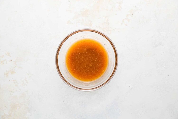 A clear glass bowl contains a small amount of light brown liquid, resembling a sauce or dressing for Holiday Brussels Sprouts, placed on a white textured surface.