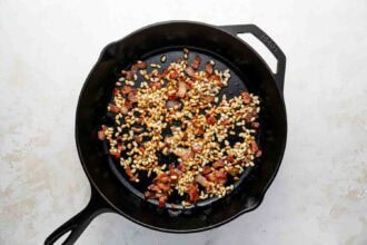 A cast iron skillet containing toasted pine nuts and cooked bacon pieces, perfect for topping Holiday Brussels Sprouts, sits on a light-colored surface.