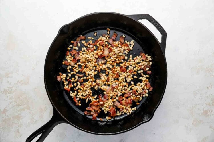 A cast iron skillet containing toasted pine nuts and cooked bacon pieces, perfect for topping Holiday Brussels Sprouts, sits on a light-colored surface.