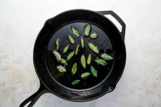 Fresh sage leaves frying in oil inside a black cast iron skillet on a light-colored surface