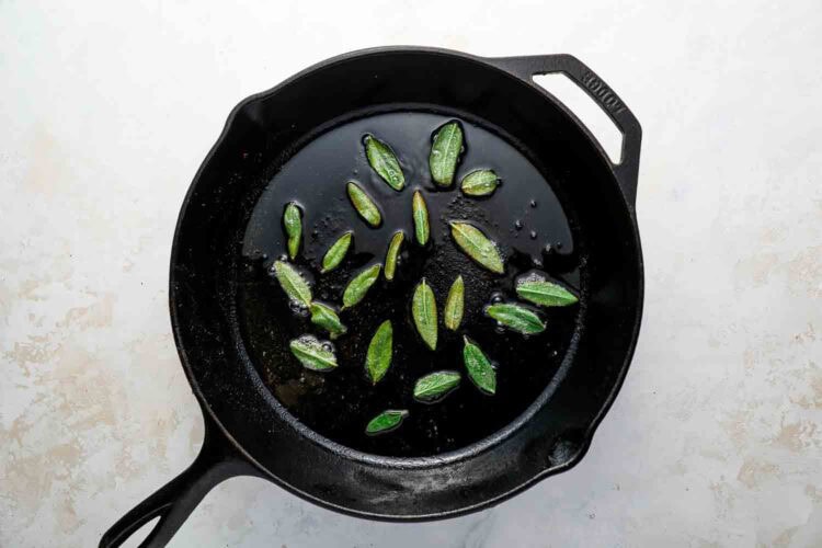 Fresh sage leaves frying in oil inside a black cast iron skillet on a light-colored surface