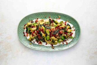 A green oval plate with Brussels Sprouts, pomegranate seeds, toasted pine nuts, herbs, and a creamy white sauce on a light background.