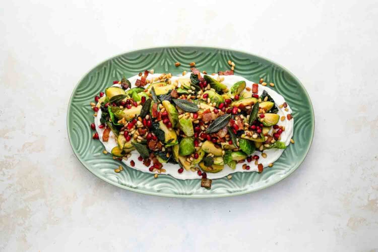 A green oval plate with Brussels Sprouts, pomegranate seeds, toasted pine nuts, herbs, and a creamy white sauce on a light background.