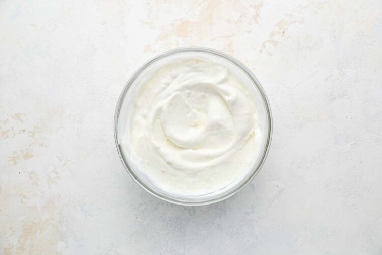 A glass bowl filled with plain, creamy yogurt sits on a light-colored surface, ready to complement your Holiday Brussels Sprouts.