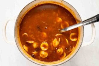 A pot of Marry Me Turkey Soup, a rich tomato-based soup with orecchiette pasta and a metal ladle, viewed from above on a white surface.
