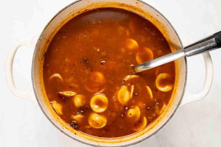 A pot of Marry Me Turkey Soup, a rich tomato-based soup with orecchiette pasta and a metal ladle, viewed from above on a white surface.