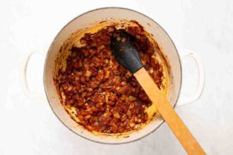 A white pot containing sautéed onions and spices for Marry Me Turkey Soup, being stirred with a black-tipped wooden spatula on a white surface.