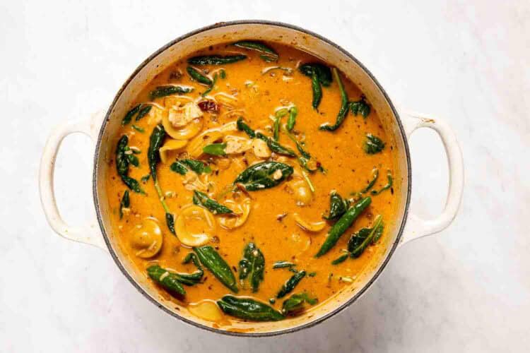 A white pot filled with creamy orange curry, featuring spinach, mushrooms, and sliced onions, resembles a cozy Marry Me Turkey Soup when viewed from above on a white surface.