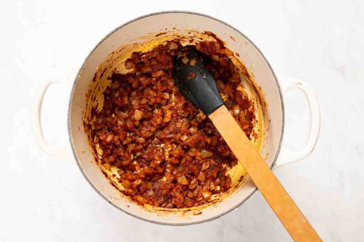 A white pot containing sautéed onions and spices for Marry Me Turkey Soup, being stirred with a black-tipped wooden spatula on a white surface.