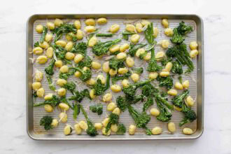 Baby potatoes, broccolini, and pillowy Sheet Pan Gnocchi are spread out on a metal baking sheet, perfectly seasoned and ready for roasting.