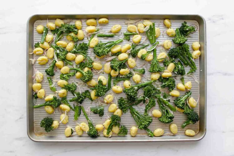 Baby potatoes, broccolini, and pillowy Sheet Pan Gnocchi are spread out on a metal baking sheet, perfectly seasoned and ready for roasting.