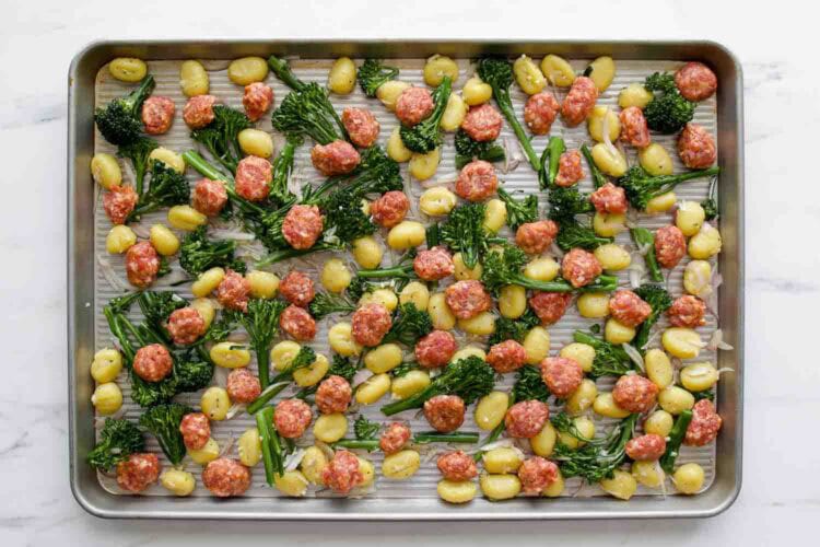 A Sheet Pan Gnocchi dinner featuring uncooked gnocchi, broccolini, and pieces of sausage evenly spread out on a baking sheet, ready for roasting.
